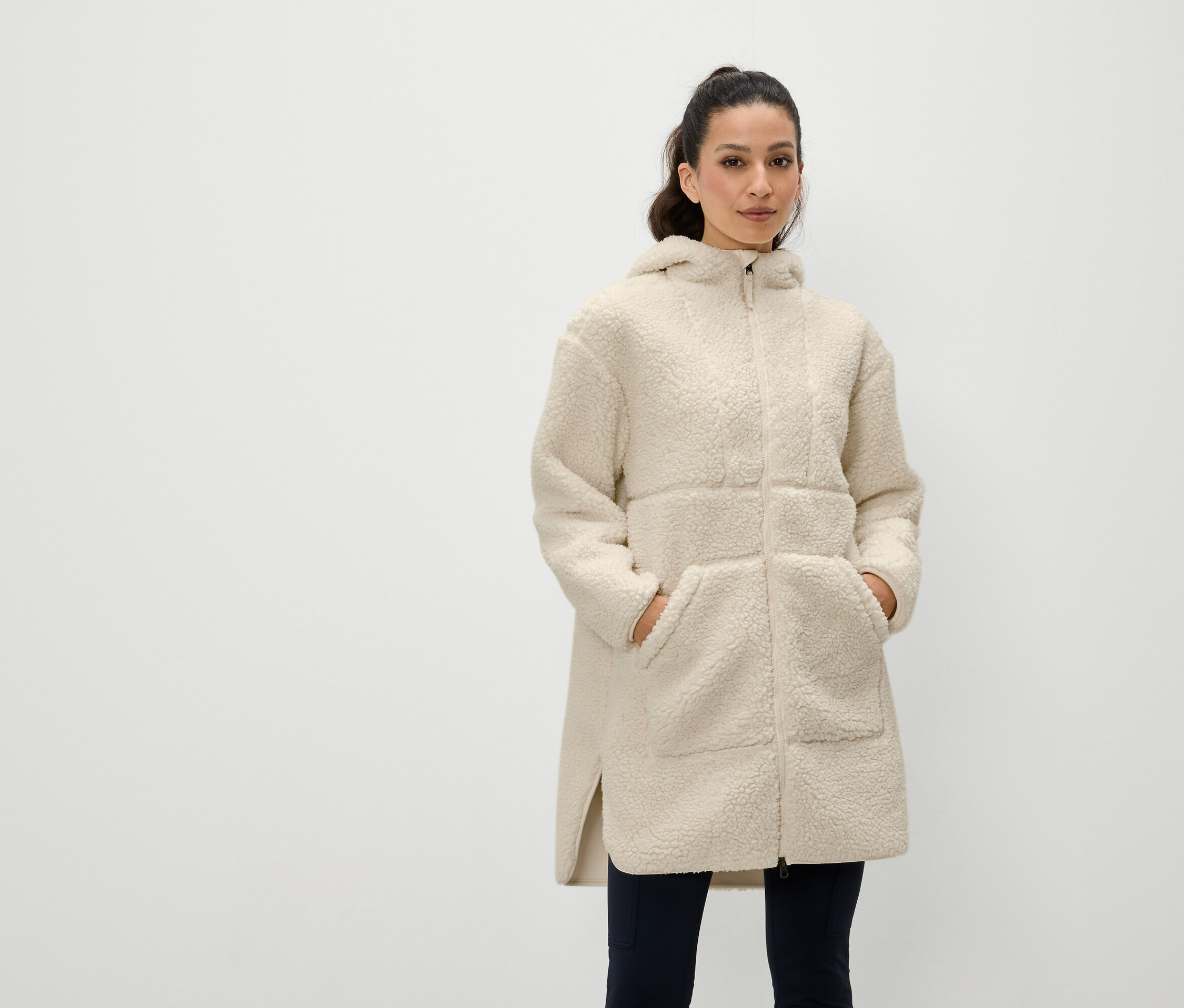 Žena stojí s rukama v kapsách krémového kabátu s kapucí na zip.