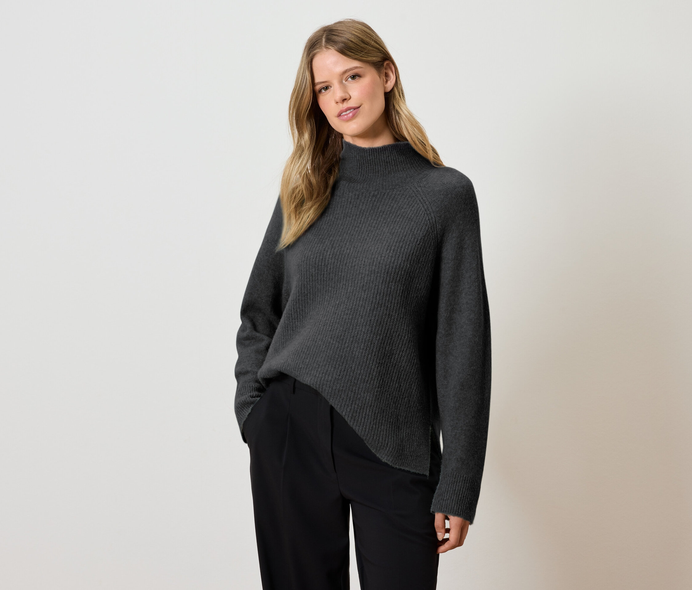 Žena pózuje v šedém svetru a černých kalhotách.