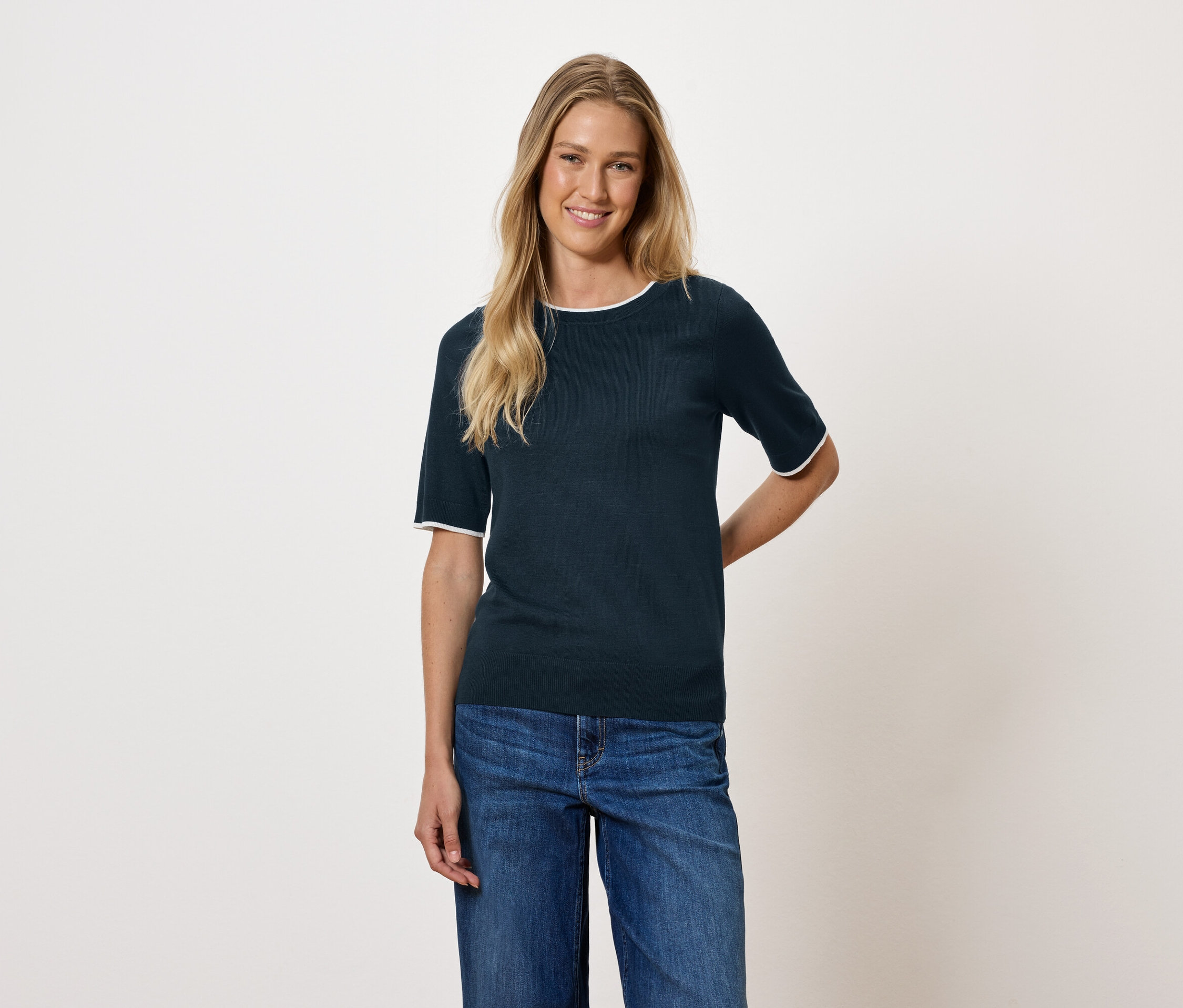 Žena s blond vlasy má na sobě tmavě modrý top s krátkým rukávem a džíny.