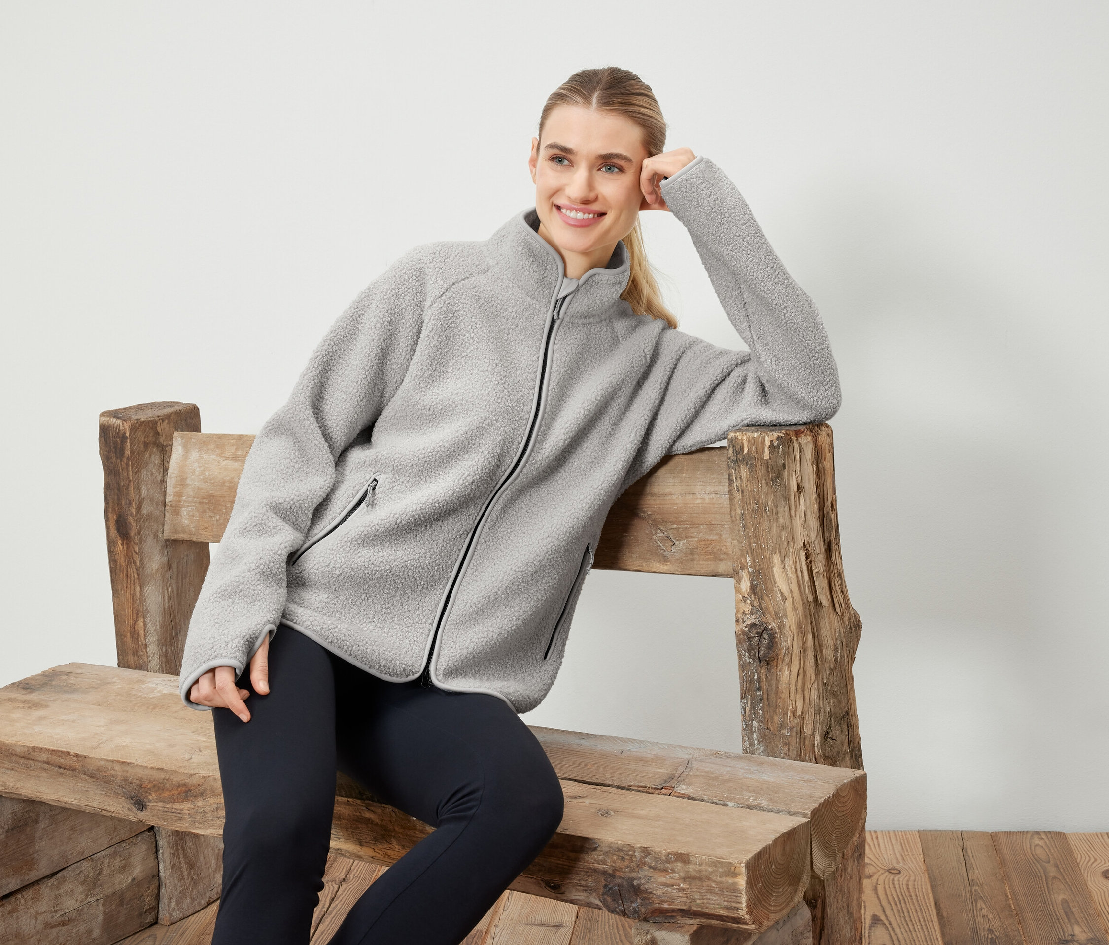 Žena v šedé fleecové bundě pózuje na dřevěné lavičce.