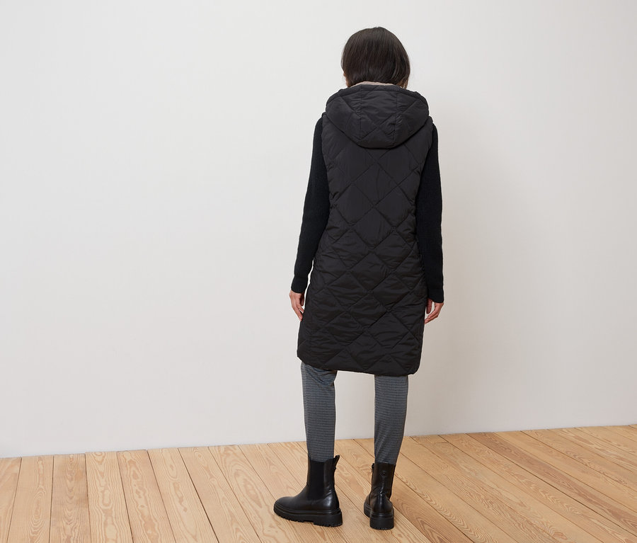 Žena zády k fotoaparátu má na sobě prošívanou černou vestu s kapucí, černé boty a šedé legíny.