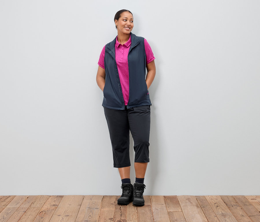Žena stojí s rukama v kapsách před zdí. Má na sobě tmavě modrou vestu přes růžové polo tričko, černé kalhoty a boty.