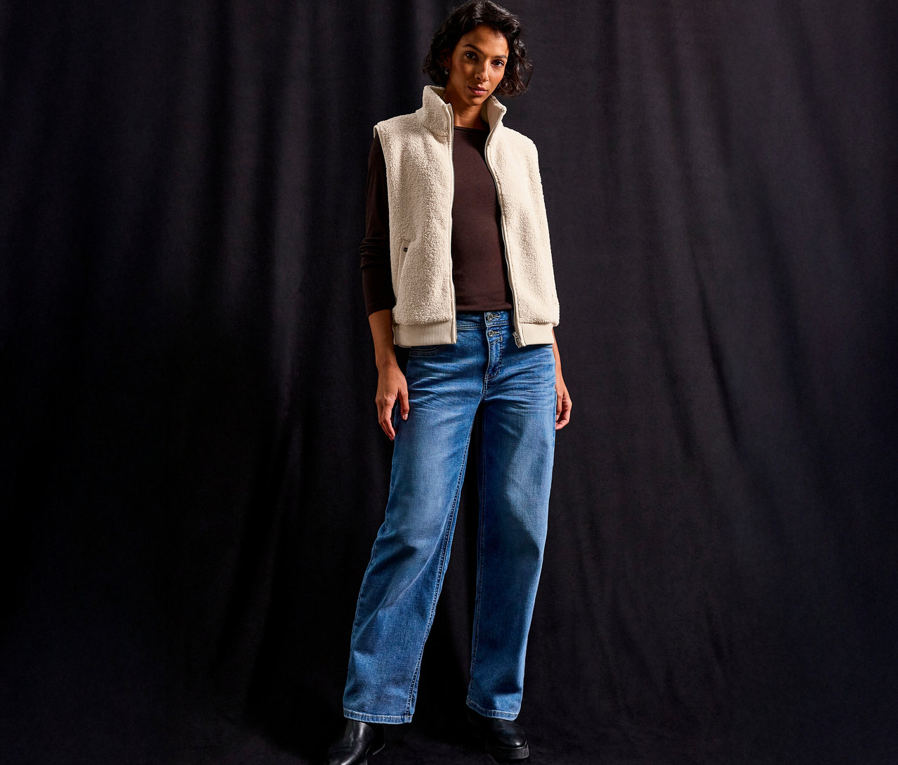 Žena má na sobě plyšovou vestu Street One přes hnědé tričko s dlouhým rukávem a džíny na černém pozadí.