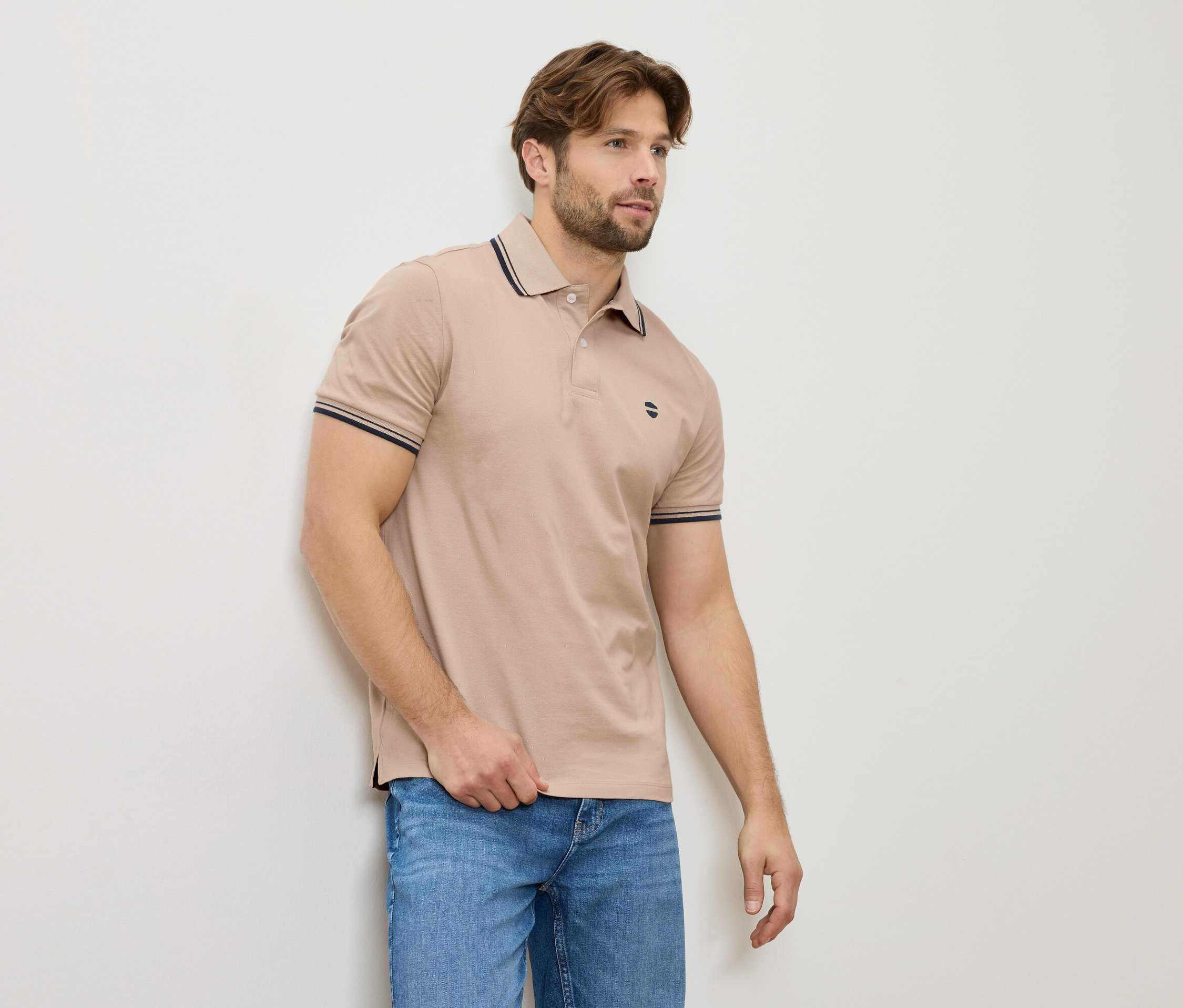 Muž v béžové polokošili a džínách stojí u zdi.