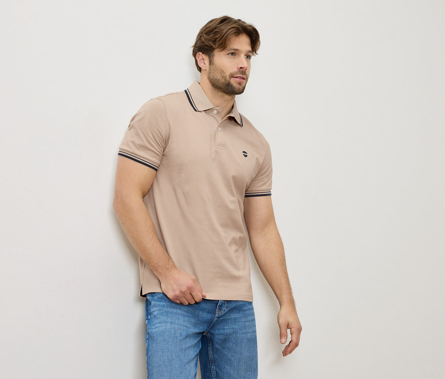 Muž v béžové polokošili a džínách stojí u zdi.