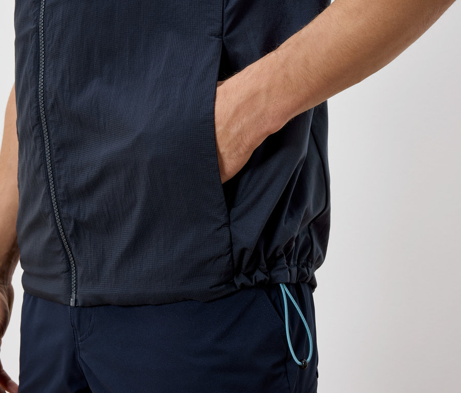 Detail osoby v tmavě modré vestě a kalhotách, s rukou v kapse vesty.