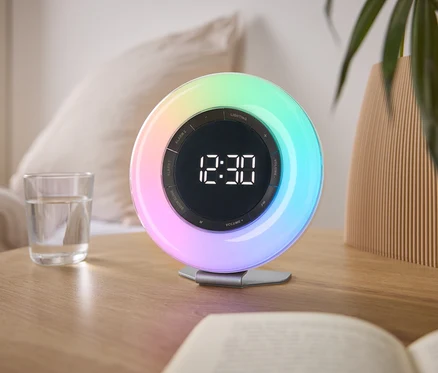 Na nočním stolku stojí barevný světelný budík ukazující čas 12:30, vedle sklenice vody a knihy.