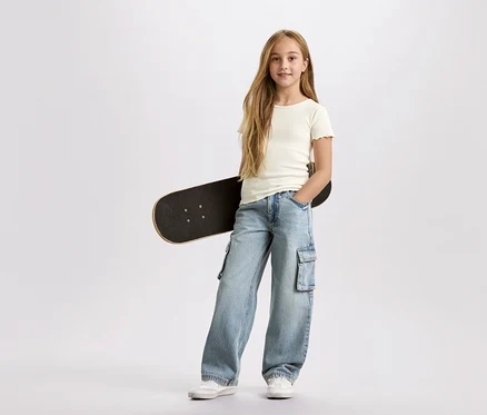 Dívka s dlouhými blond vlasy, s černým skateboardem v podpaží, má na sobě světle modré cargo kalhoty a bílé tričko na bílém pozadí.