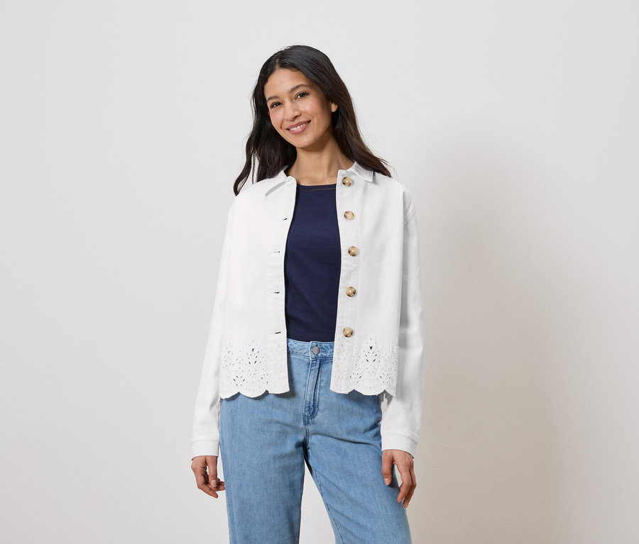 Žena má na sobě bílou bundu s dírkovaným vzorem přes tmavě modrý top a světle modré džíny.