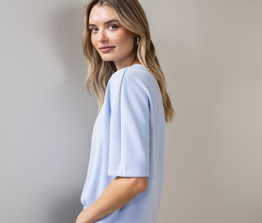 Žena se dívá přes rameno, má na sobě světle modré triko Street One s krátkými rukávy a stříbrné kruhové náušnice.
