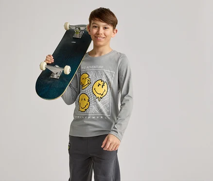 Usmívající se chlapec drží skateboard na rameni, má na sobě šedé tričko s dlouhým rukávem se žlutými motivy smajlíků a šedé kalhoty.