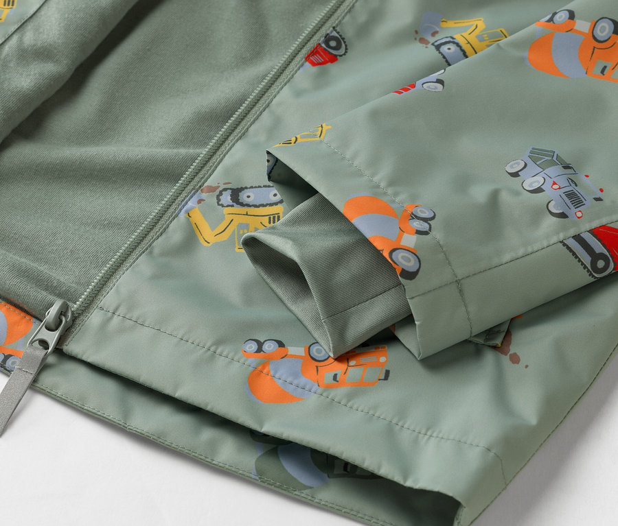 Detailní záběr na olivově zelenou bundu se vzorem stavebních vozidel, částečně rozepnutou na zip.