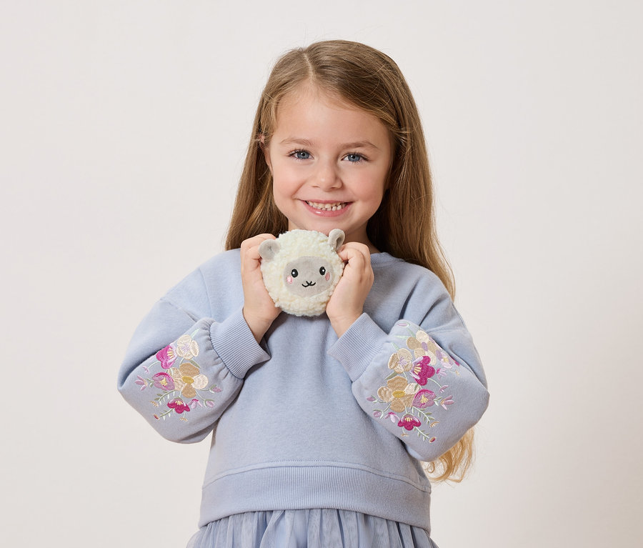 Usmívající se dívka drží před sebou plyšovou ovečku. Má na sobě modrý svetr s květinovými výšivkami na rukávech.