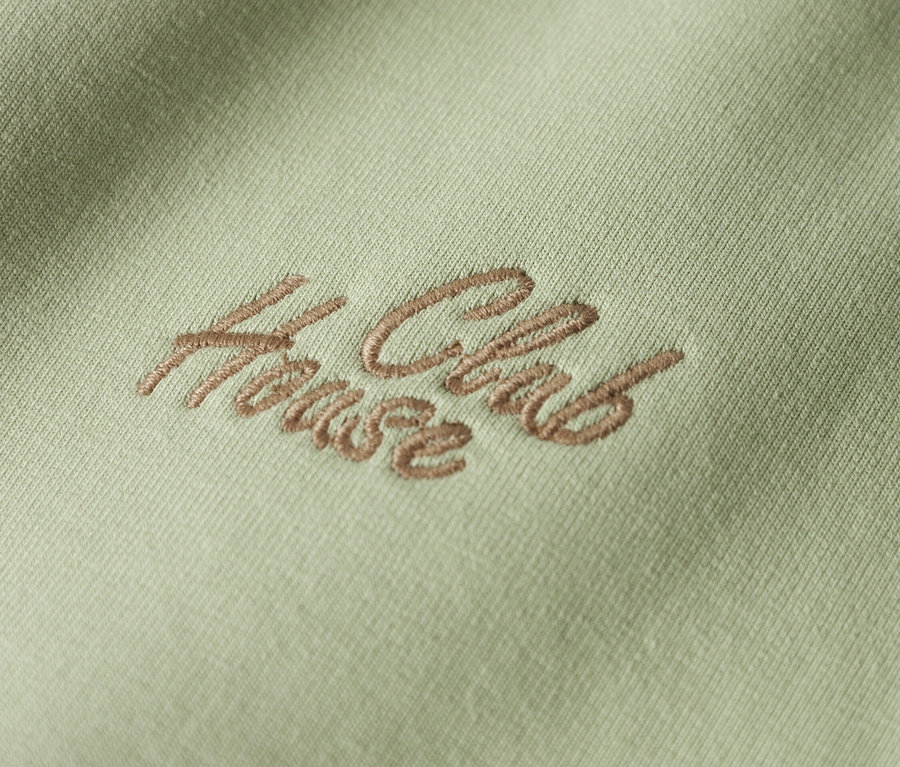 Detailní záběr světle zelené látky s hnědou výšivkou "House Club".