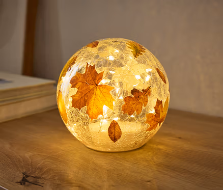 Kulatá lampa s prasklou skleněnou texturou, zdobená podzimními listy, teple svítí na dřevěném povrchu. Malá světla uvnitř osvětlují listy.
