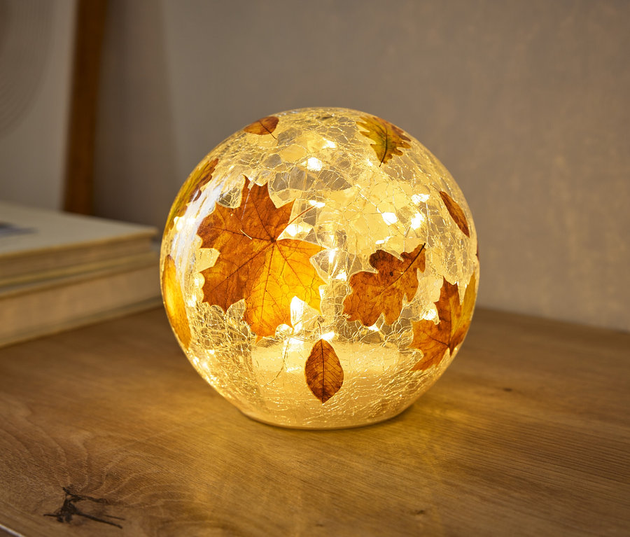 Kulatá lampa s prasklou skleněnou texturou, zdobená podzimními listy, teple svítí na dřevěném povrchu. Malá světla uvnitř osvětlují listy.