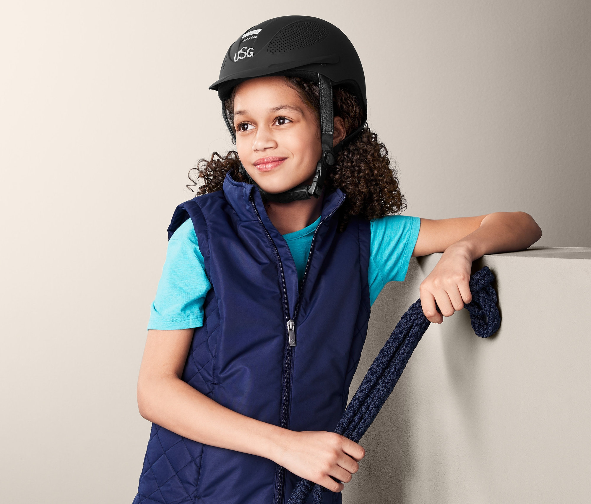 Dívka s hnědými kudrnatými vlasy má na sobě modrou jezdeckou vestu a černou dětskou jezdeckou helmu USG a drží modré lano.