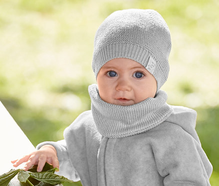 Kojenecká dívka stojí u stolu. Má na sobě pletenou čepici a šálu, fleecovou bundu s recyklovaného materiálu a pohodlné kalhoty.