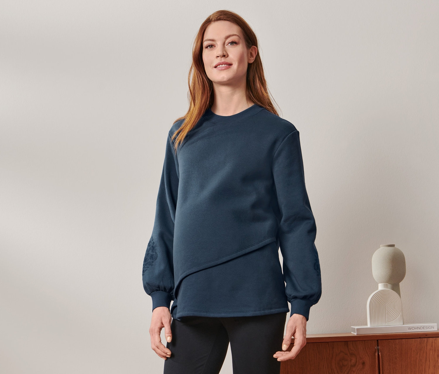 Těhotná žena stojí a má na sobě těhotenskou mikinu, černé kalhoty a tenisky.