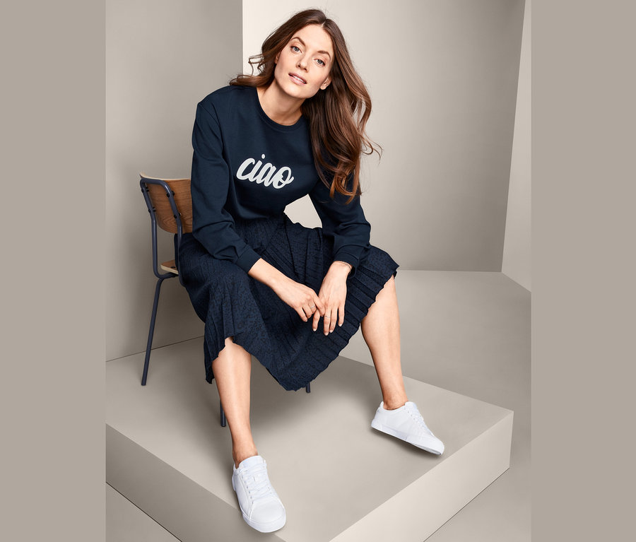 Žena sedí na židli a má na sobě tmavě modrou mikinu s nápisem a bílé tenisky.