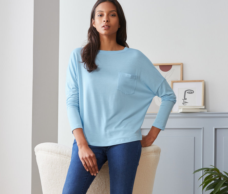 Žena pózuje v modrém tričku s netopýřími rukávy, džínách a bílých teniskách.