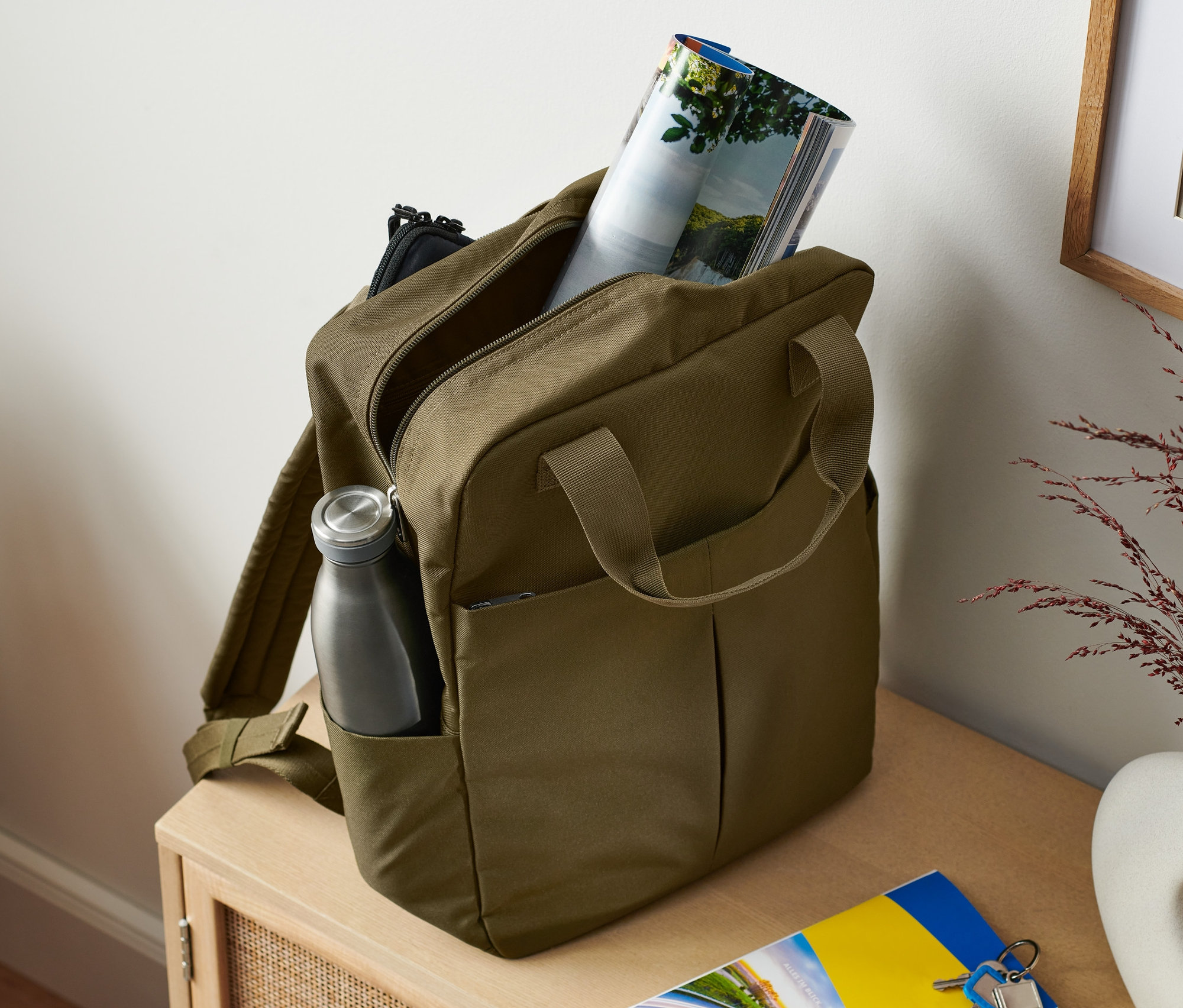 Khaki bezpečnostní batoh s lahví na vodu v boční kapse a časopisem uvnitř.