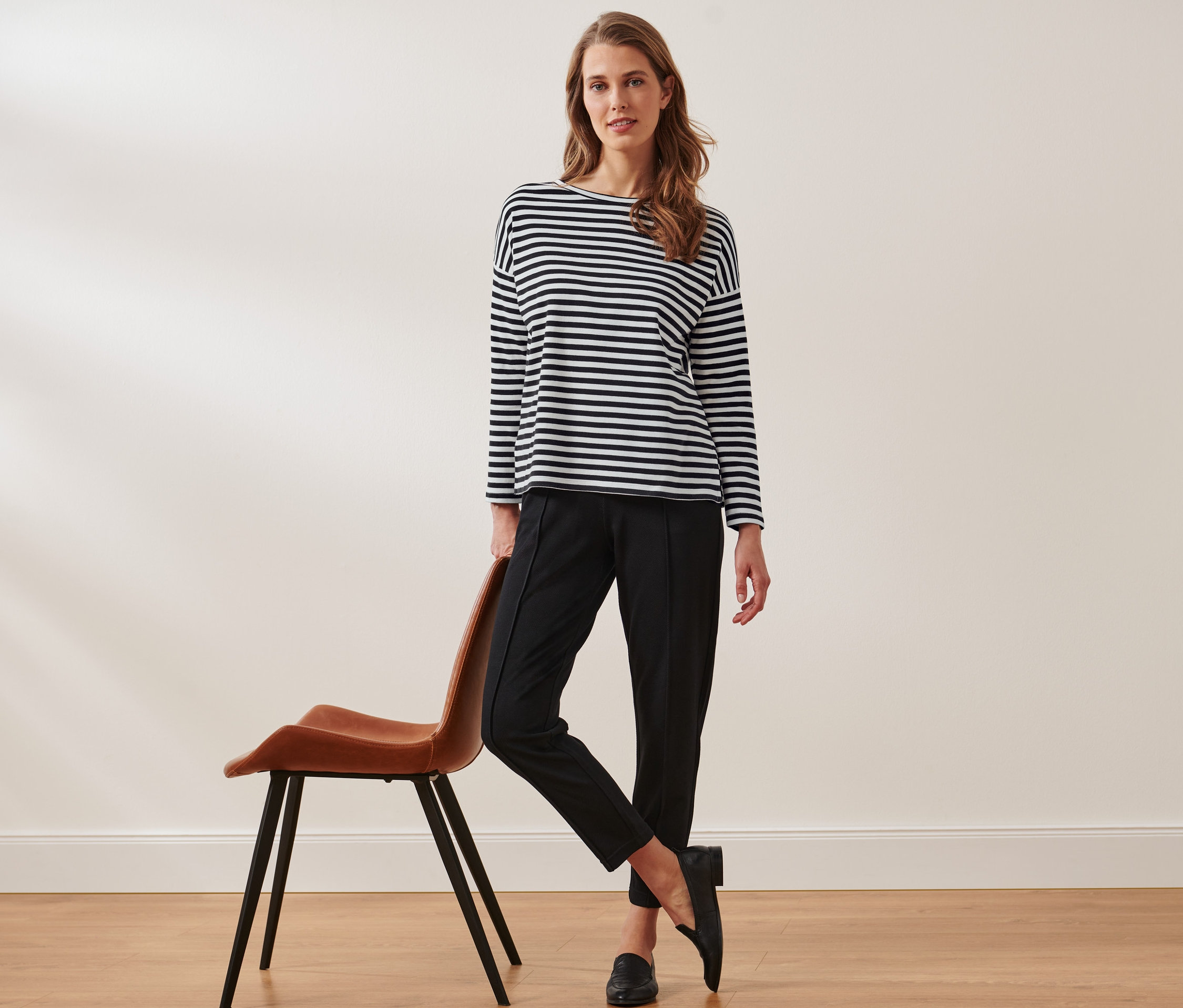 Žena opřená o hnědou židli pózuje v černých kalhotách a pruhovaném tričku s dlouhým rukávem.