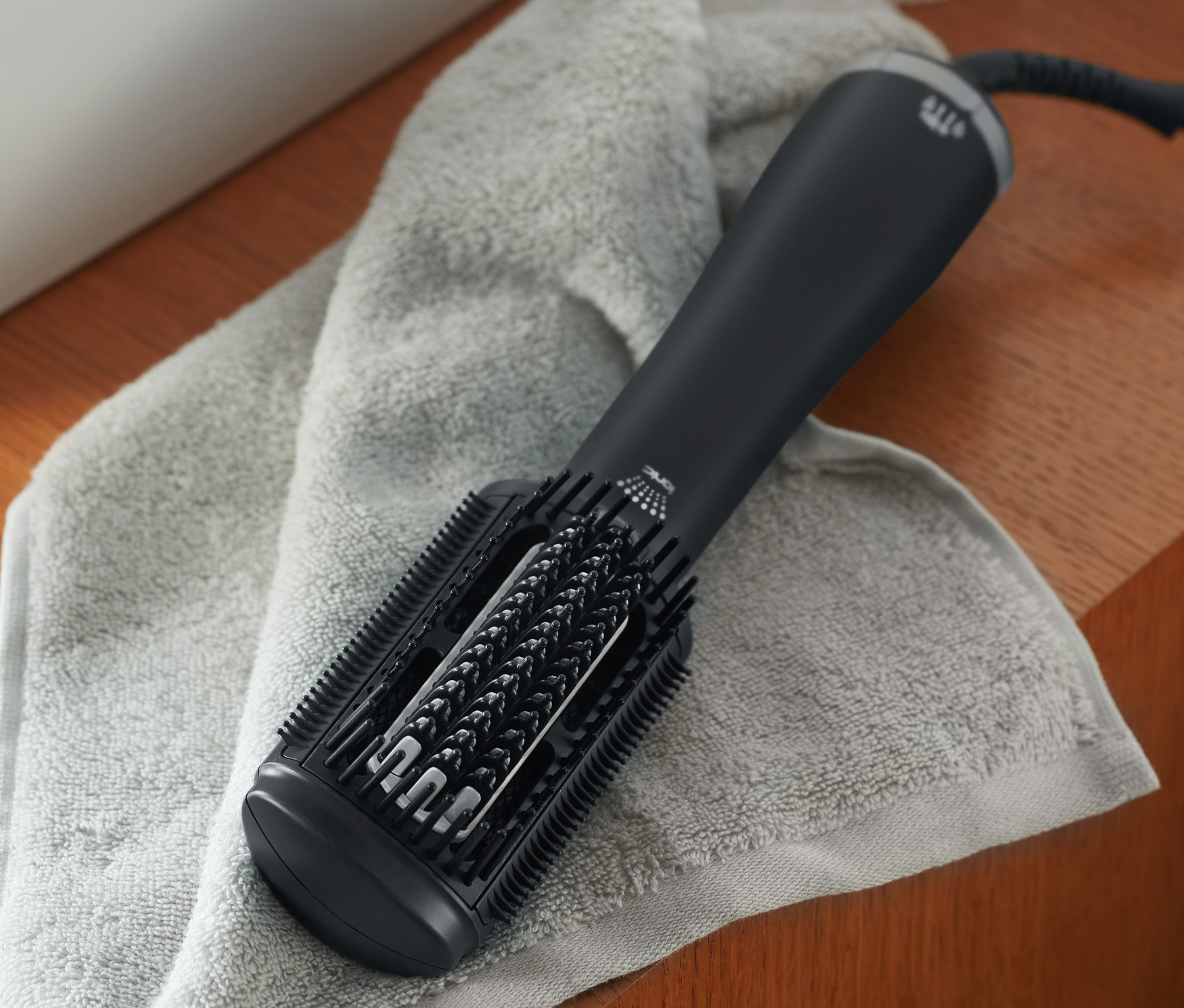 Černý žehlicí kartáč na vlasy Premium leží na šedé osušce vedle bílého umyvadla a dávkovače mýdla.