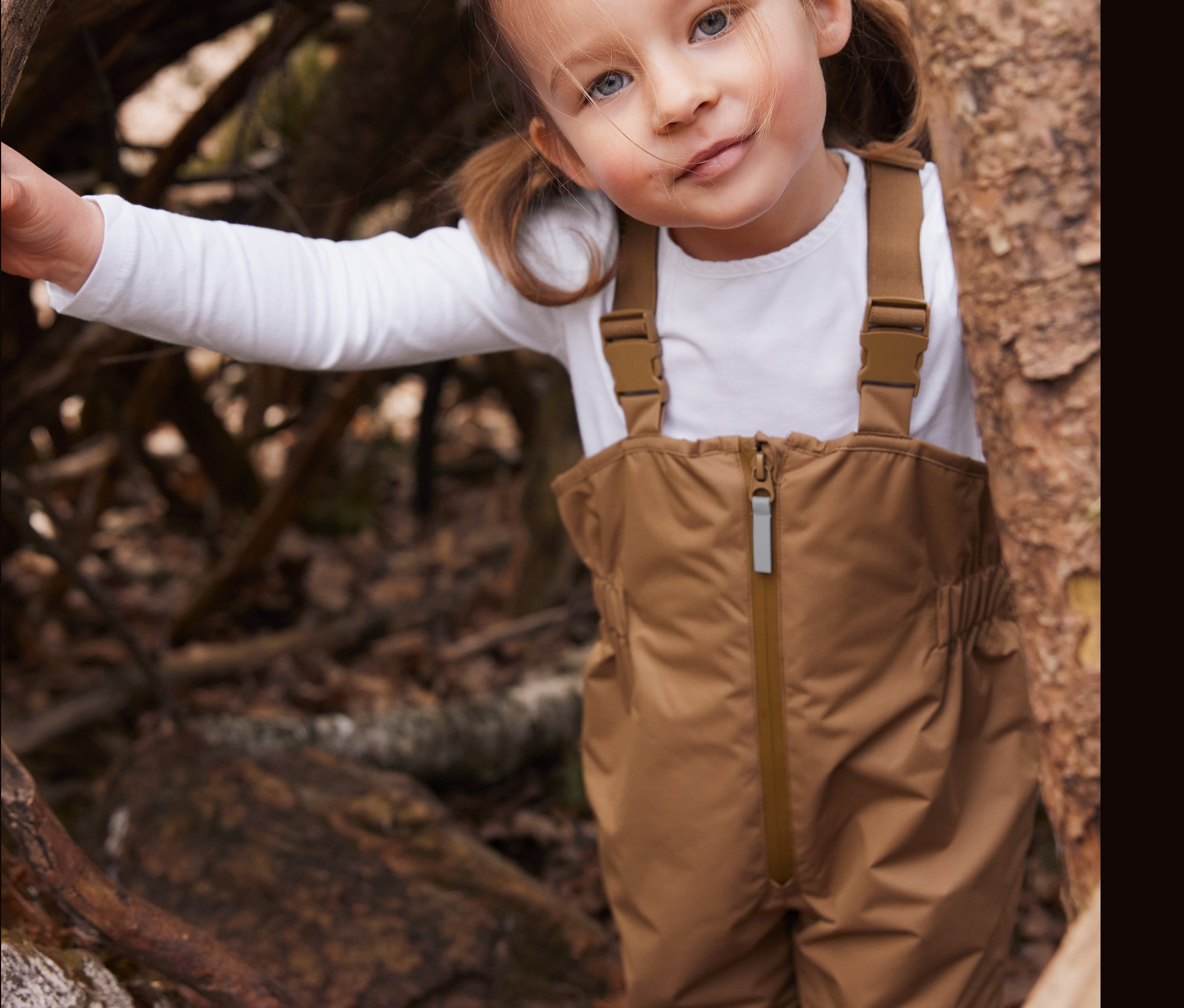 Dívka v hnědých kalhotách do deště stojí u stromu.