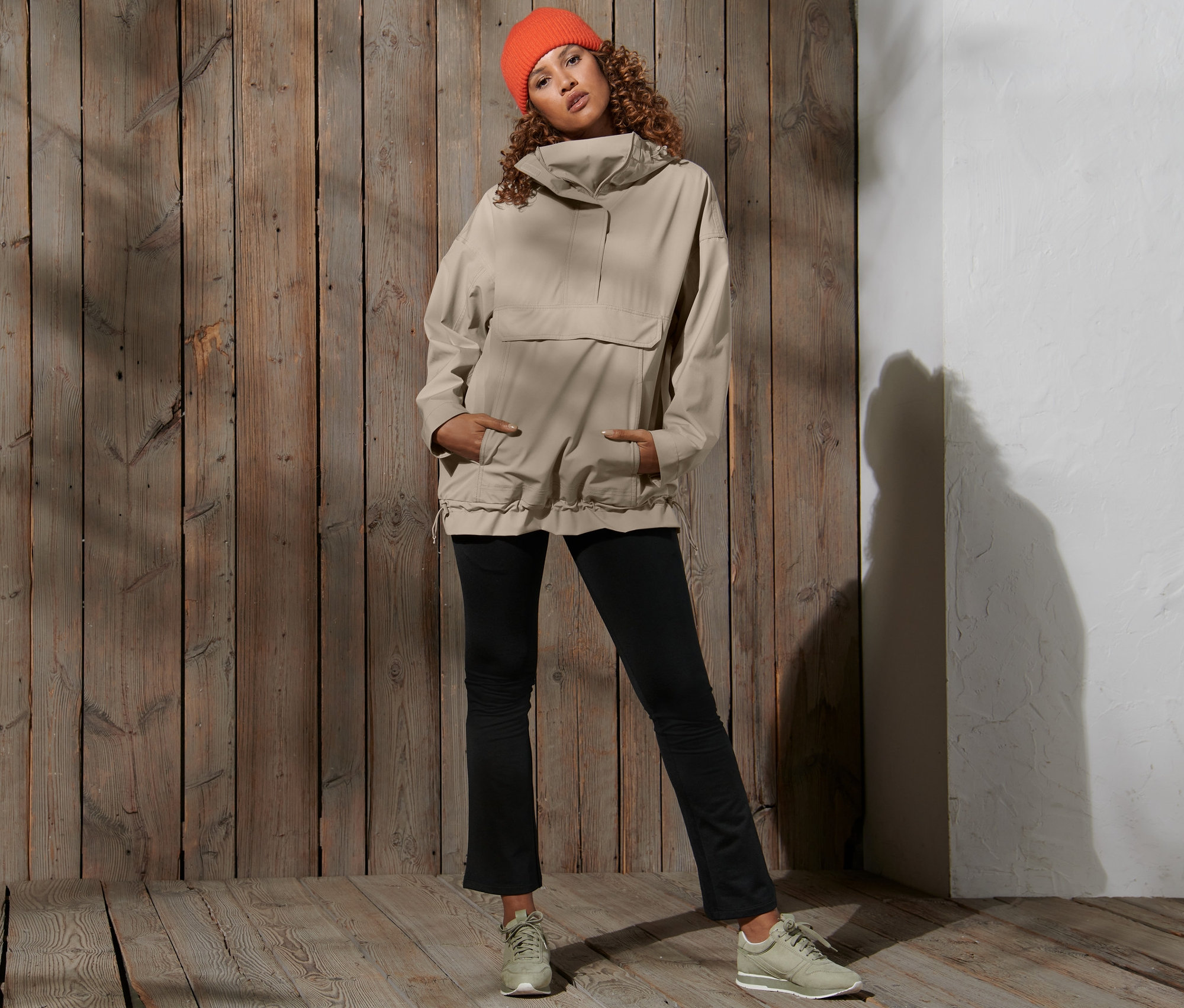 Žena pózuje na dřevěném pozadí a má na sobě oranžovou čepici a béžovou oversized funkční bundu do deště.