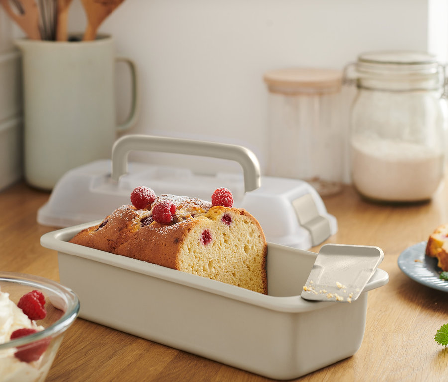 Nakrájený koláč ve hranaté formě s přepravním víkem, posypaný moučkovým cukrem a malinami.