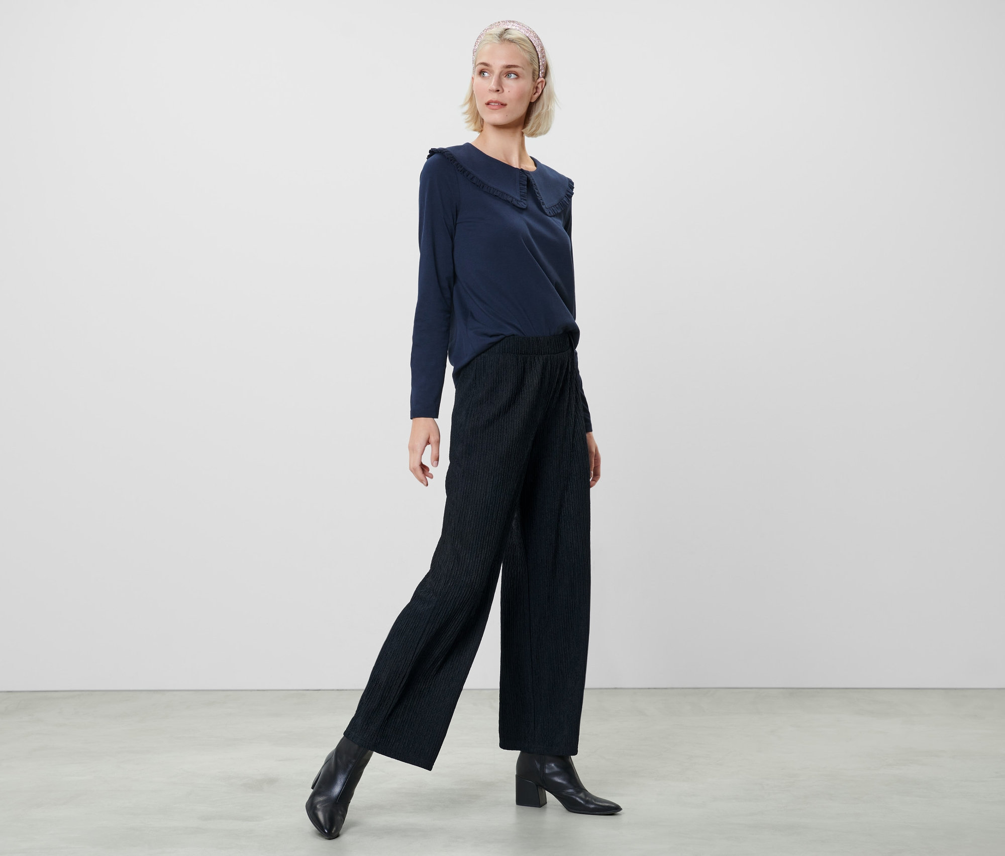 Žena pózuje v tmavě modré halence a tmavě modrých plisovaných kalhotách.