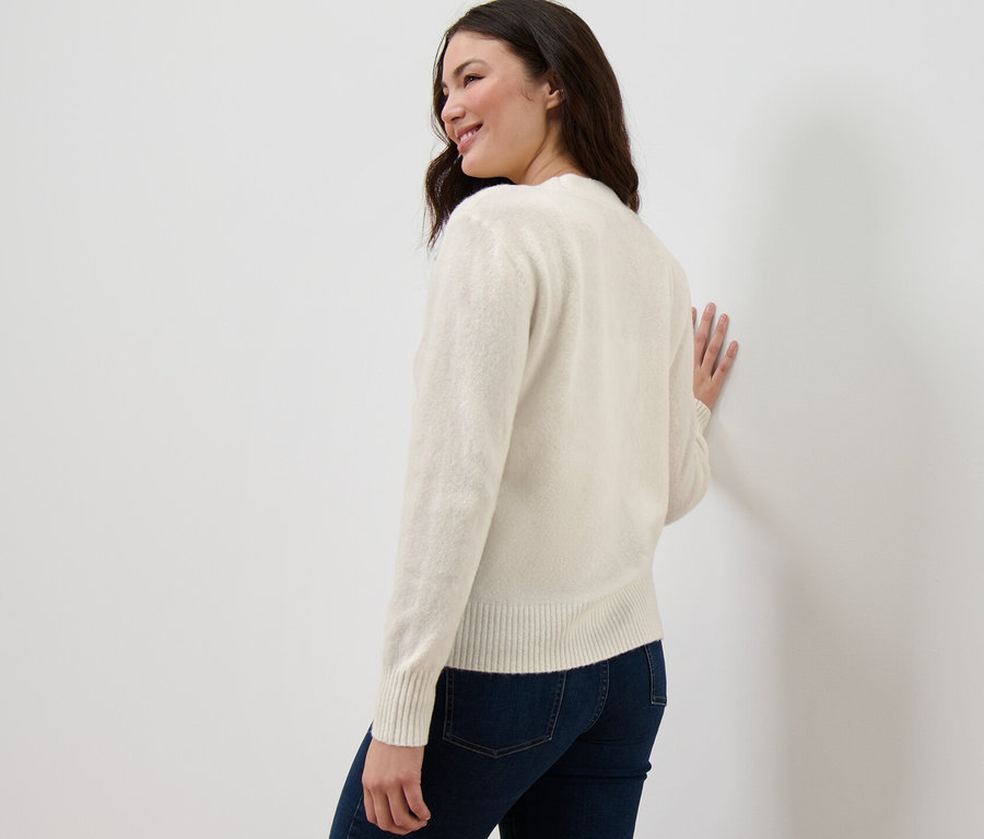 Žena se dívá přes rameno a opírá se o zeď v krémově bílém kardiganu s pletenou strukturou.