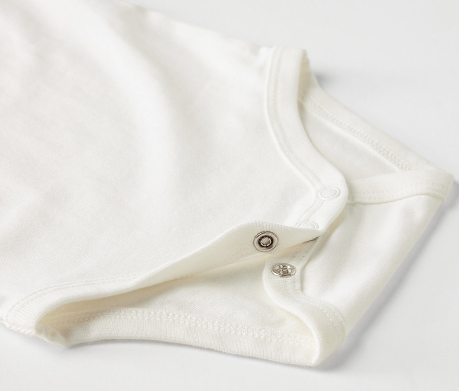 Detail bílého kojeneckého body s krátkým rukávem a patentkami.