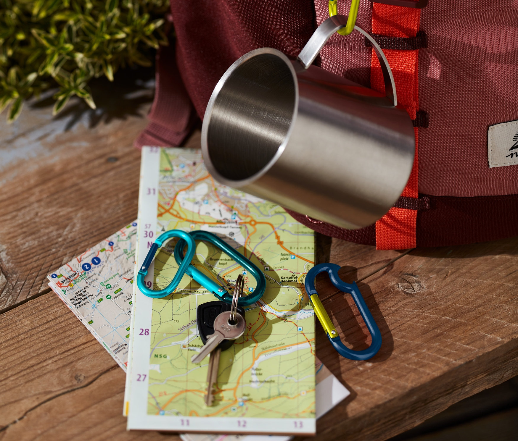 Nerezový hrnek visí na batohu pomocí jedné ze 4 karabin. Na mapě leží klíče s další karabinou.