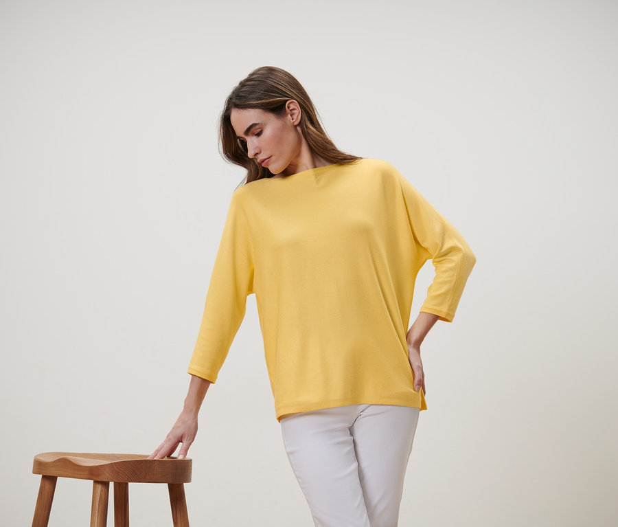 Žena se sklání k dřevěné stoličce, má na sobě žlutý svetr z jemného úpletu a bílé kalhoty.