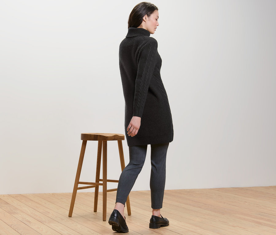 Žena odvrácená zády, má na sobě dlouhý černý pletený svetr s copánkovým vzorem.