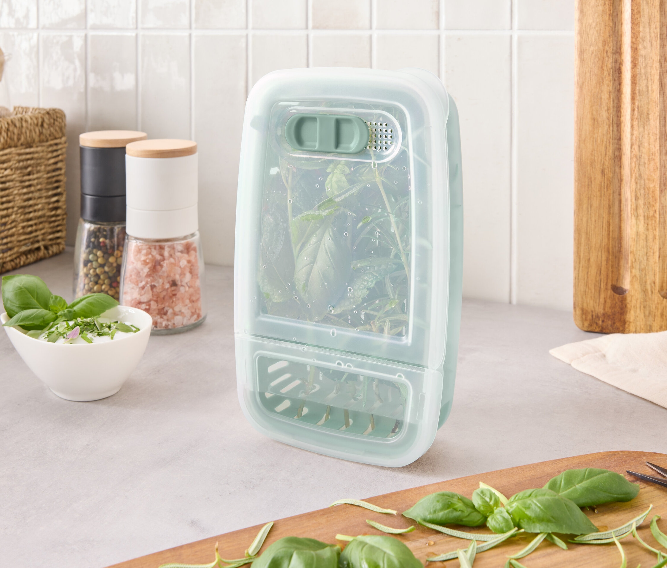 Nádoba na uchovávání bylinek s bylinkami uvnitř stojí na lince vedle ingrediencí a prkénka.