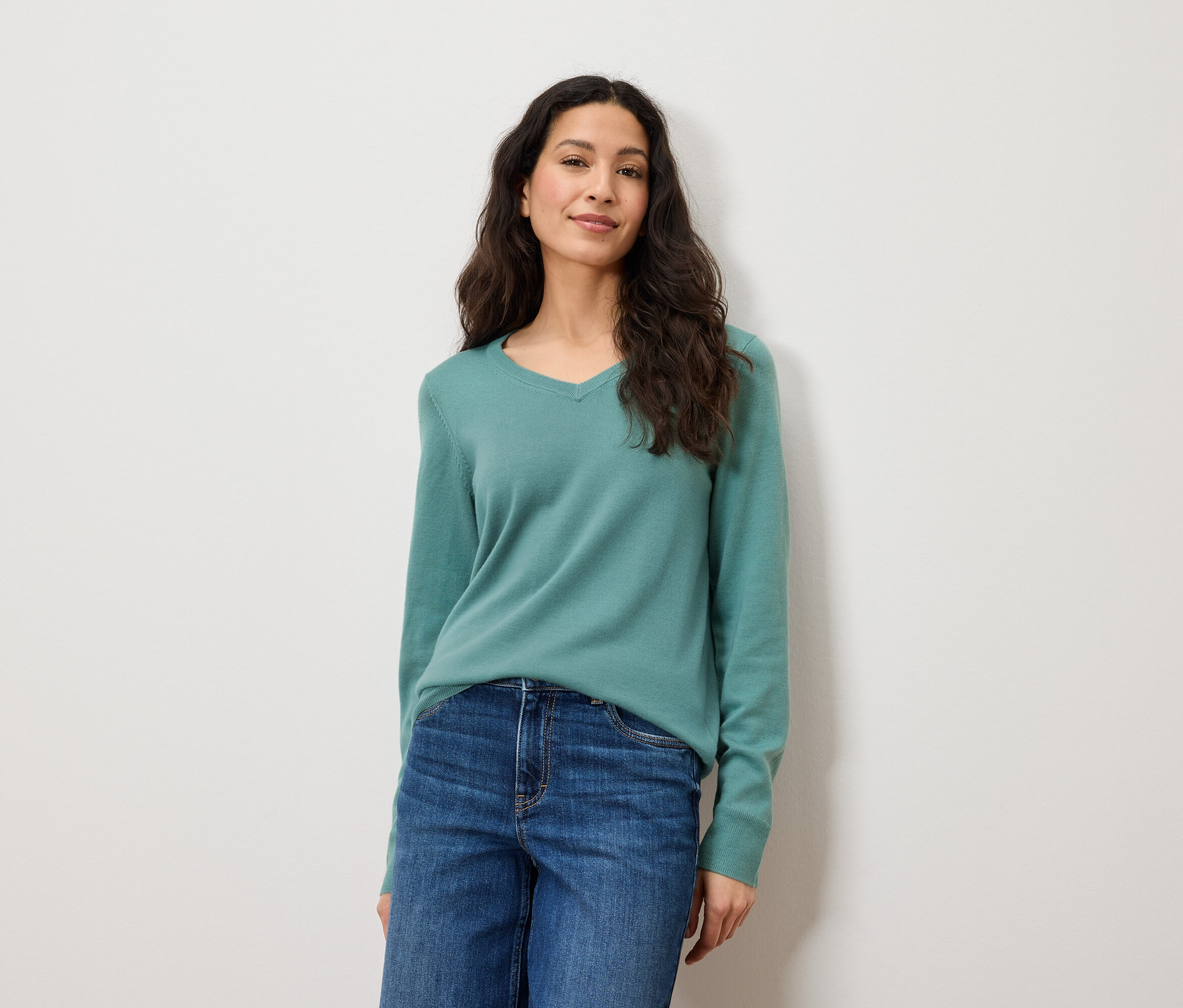 Žena s dlouhými hnědými vlasy pózuje na bílém pozadí. Má na sobě modré džíny a zelený svetr.