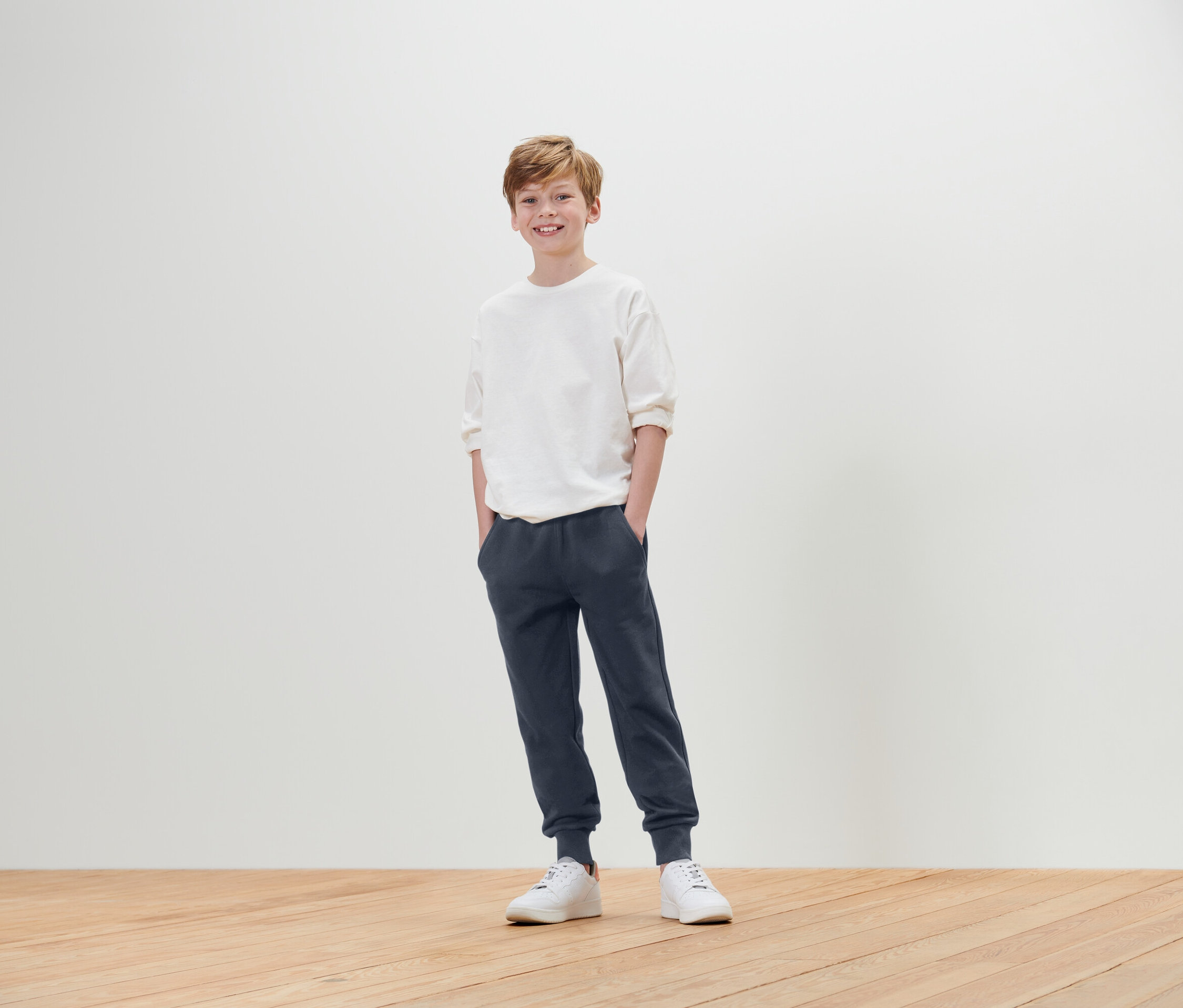 Chlapec stojí s rukama v kapsách a má na sobě bílé tričko, modré tepláky a bílé tenisky.