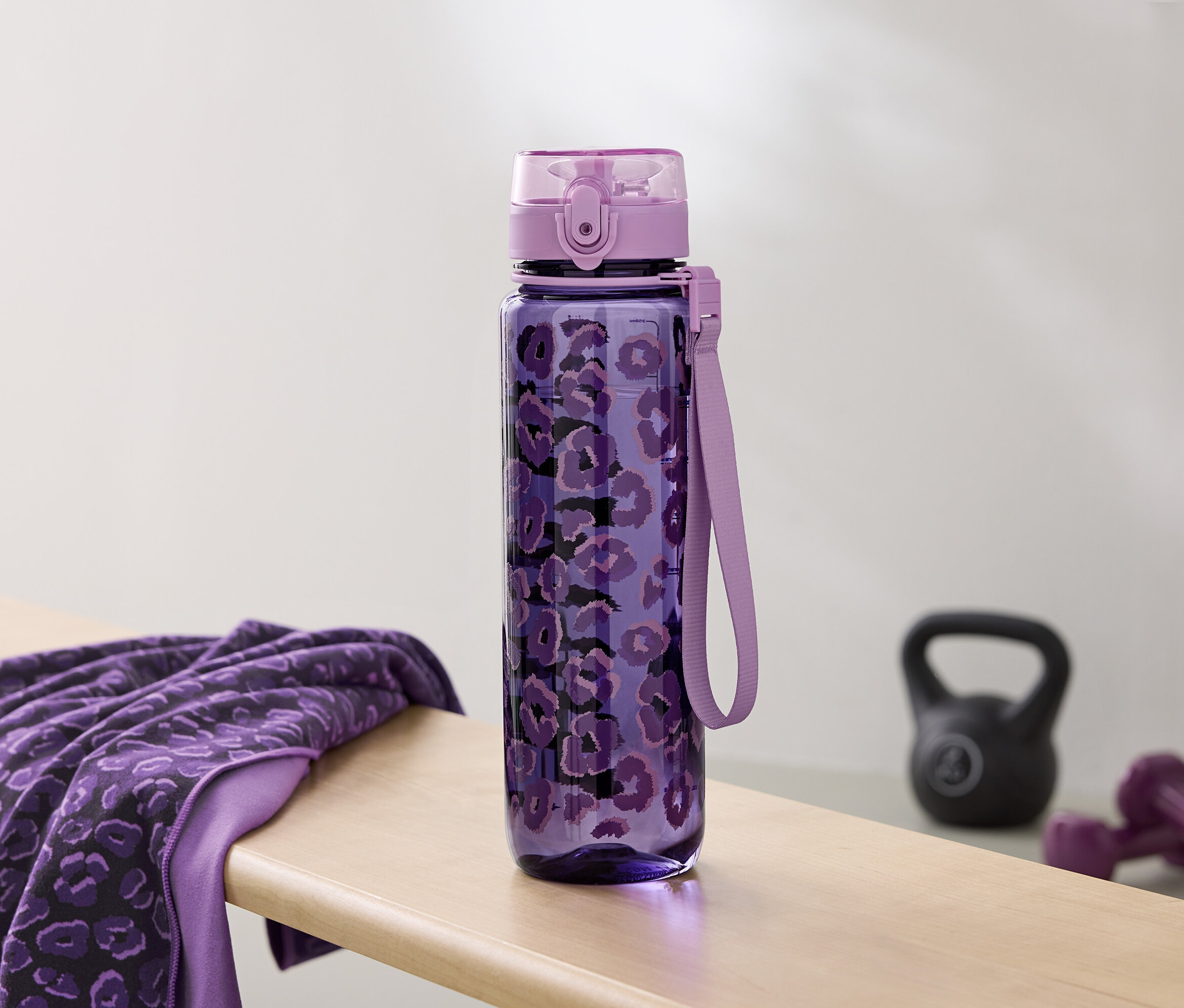Fialová láhev na vodu s potiskem leží vedle ručníku a činek na lavici.