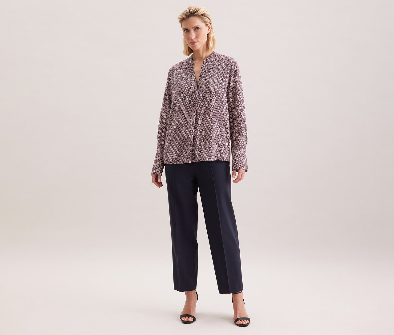 Žena má na sobě tuniku z viskózy Seidensticker s celoplošným potiskem v ecru a burgundské barvě, v kombinaci s tmavými kalhotami a černými sandály.