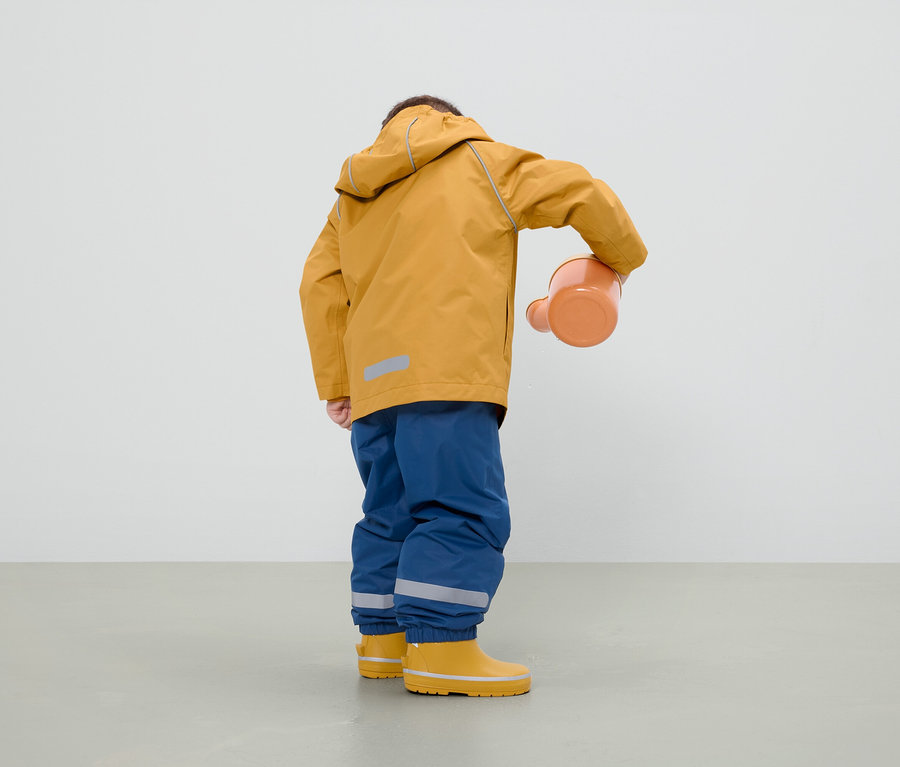 Chlapec v hořčicové bundě, modrých kalhotách a žlutých holínkách drží oranžovou konev.