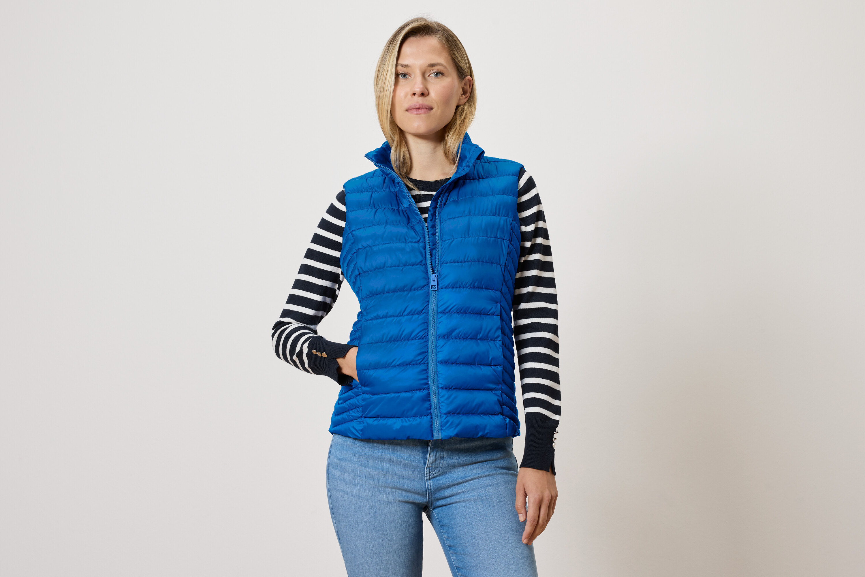 Žena v modré prošívané vestě se zipem, svetru s černými a bílými pruhy a modrých džínách.