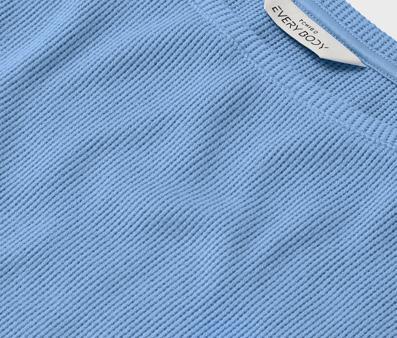 Detail světle modré textilie s žebrovanou strukturou a bílou visačkou s nápisem EVERYBODY.