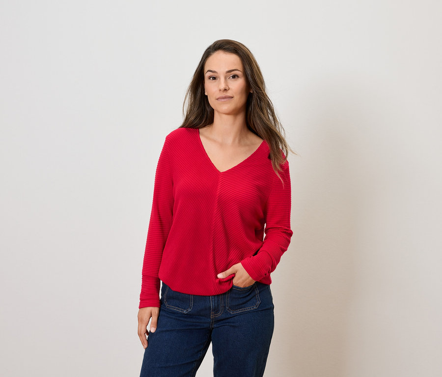 Žena s hnědými vlasy pózuje v červeném svetru s dlouhým rukávem a modrých džínách.