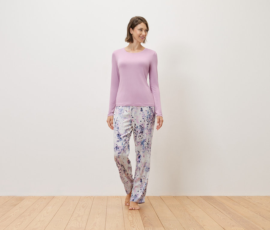 Žena stojí bosá na dřevěné podlaze, má na sobě fialový svetr a vzorované kalhoty.