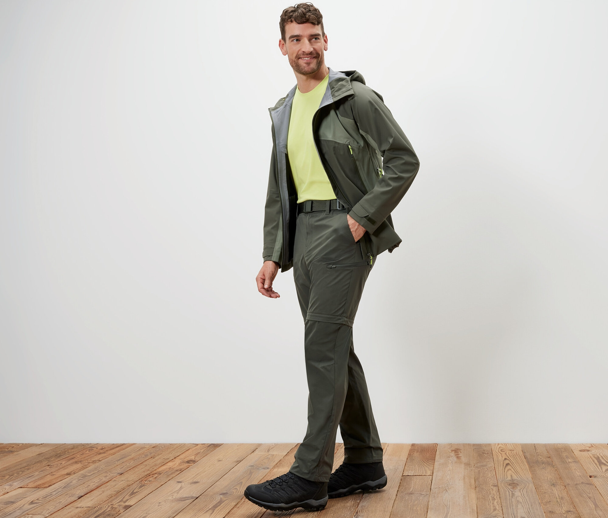 Muž pózuje v khaki 2 v 1 funkčních kalhotách a bundě na dřevěné podlaze.