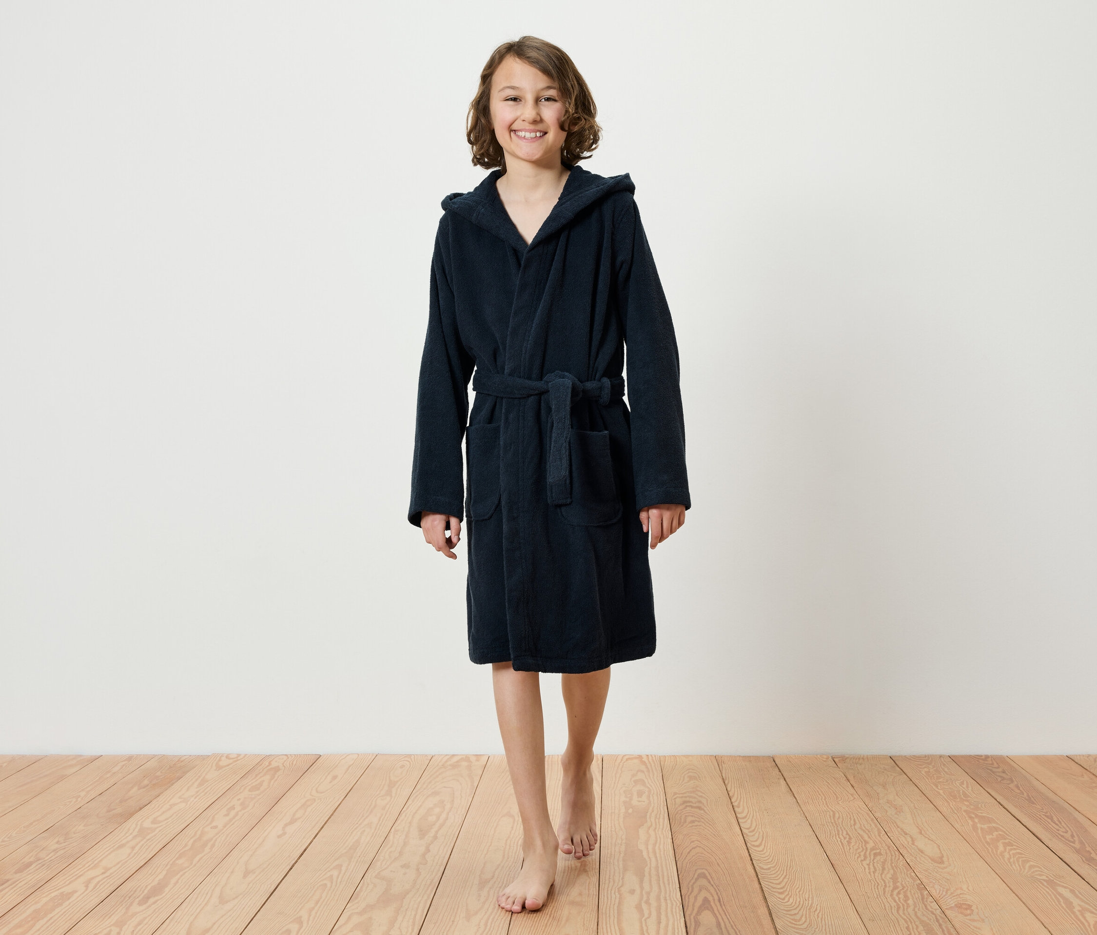 Usmívající se dospívající dívka kráčí naboso po dřevěné podlaze a má na sobě tmavě modrý župan s kapucí.