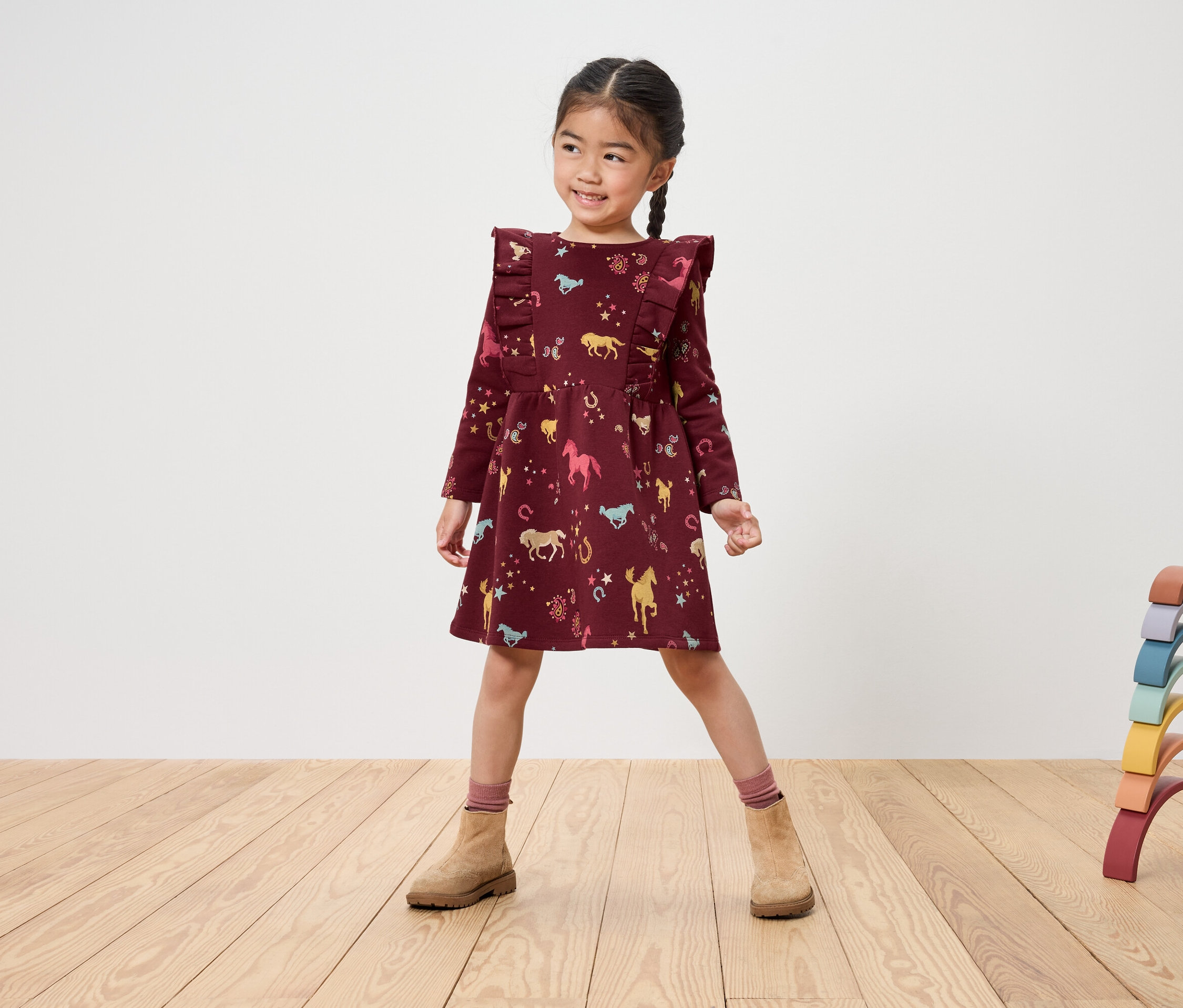 Usmívající se dívka v bordó šatech s potiskem koní a hnědých botách stojí na dřevěné podlaze vedle duhové dřevěné hračky.
